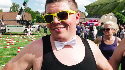 Ein Mann mit geld umrandeter Sonnenbrille, schwarzem Tanktop und weißer Fliege lacht in die Kamera