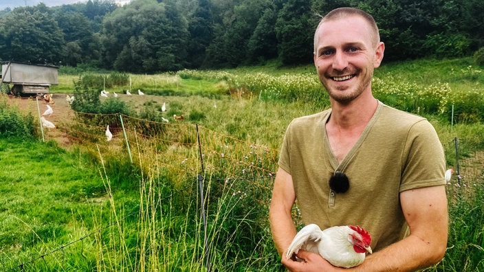 Lucas Wilhelm hält ein weißes Huhn in der Armen. Hinter ihm ist eine Wiese mit weiteren Hühnern