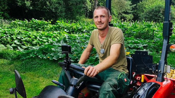 Lucas Wilhelm, olives T-Shirt, grüne Hose, kurz geschorene Haare, sitzt auf einem roten Traktor, im Hintergrund seiht man ein grünes Gemüsefeld