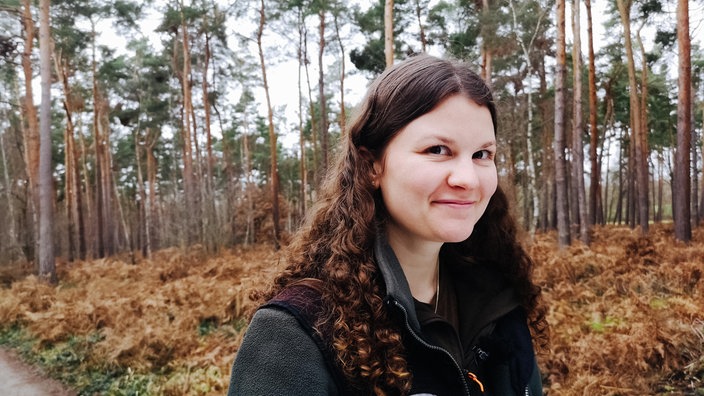 Sina Dünzer, eine junge Frau mit braunen, lockigen Haaren, steht in einem Wald und lächelt in die Kamera.