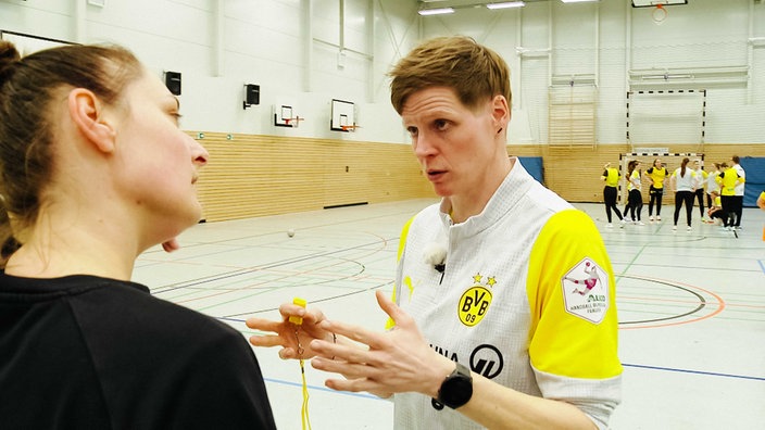 Clara Woltering steht in einer Sporthalle, in der linken Hand eine gelbe Pfeife, sie spricht mit einer Spielerin, die ihr gegenüber steht, rechts im Hintergrund stehen andere Spielerinnen vor einem Tor