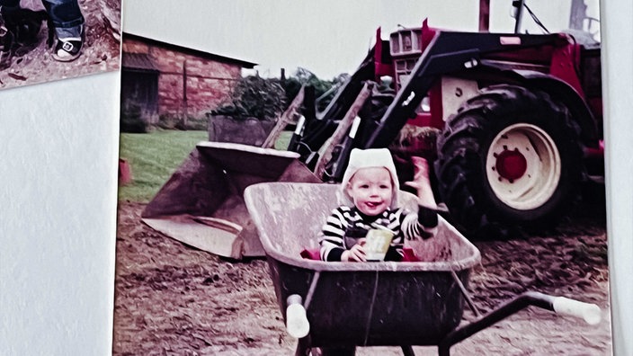 Abgefilmtes Foto von Clara Woltering als Kleinkind, das in einer Schubkarre sitzt, dahinter steht ein roter Traktor