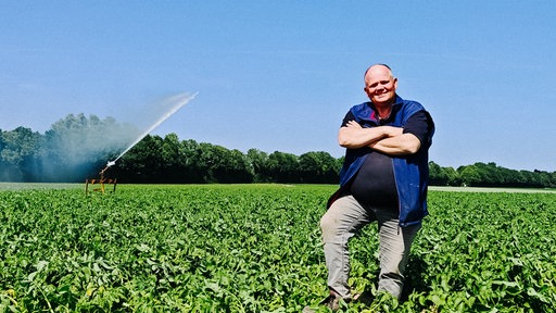 Landwirt Heinz-Georg Olligs steht auf einem grünen Feld und lächelt in die Kamera. Im Hintergrund arbeitet eine Bewässerungsmaschine.