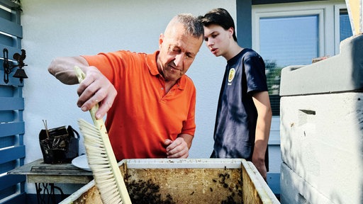 Ein älterer Mann und ein Jugendlicher stehen hinter einem Bienenstock.