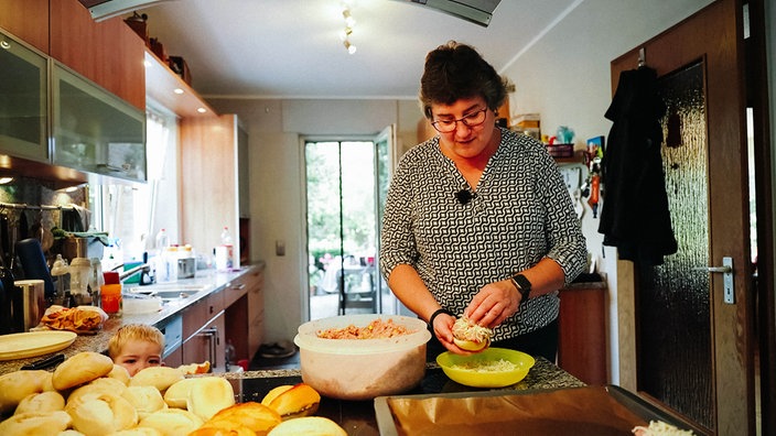 Christa Krebbing steht in der Küche und bestreut eine Brötchenhälfte mit Käse. Neben ihr steht eine große Schale mit der Hack-Mischung, daneben liegen weitere Brötchen.