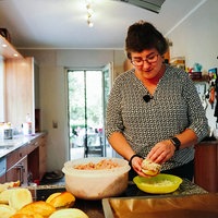 Christa Krebbing steht in der Küche und bestreut eine Brötchenhälfte mit Käse. Neben ihr steht eine große Schale mit der Hack-Mischung, daneben liegen weitere Brötchen.
