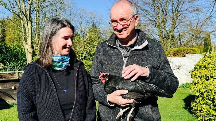 Eine Frau und ein Mann stehen nebeneinander, er hält ein schwarzes Huhn auf dem Arm