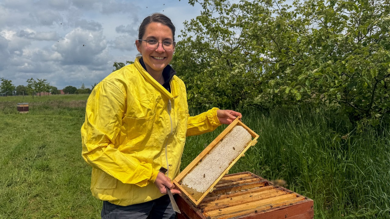 Berufsimkerin Iris van den Bongard steht in gelber Schutzjacke an einem Bienenstock, in den Händen eine Wabe