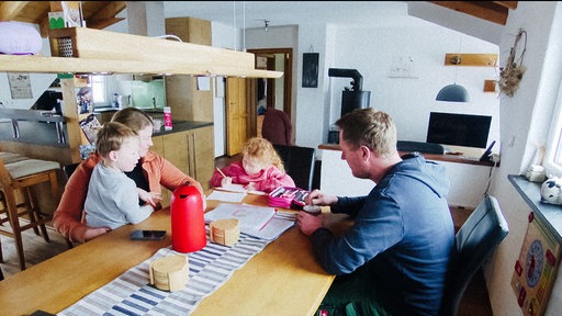 Eine vierköpfige Familie sitzt gemeinsam am Esstisch. Sohn Michel sitzt auf dem Schoß seiner Mutter Katrin Engels.Tochter Ellie ist über ihre HAusaufgaben gebeugt. Ehemann und Vater Martin Engels sitzt neben seiner Tochter-