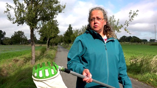 Claudia Krol steht mit einem Apfelpflücker in der Hand vor einer Straße mit Bäumen