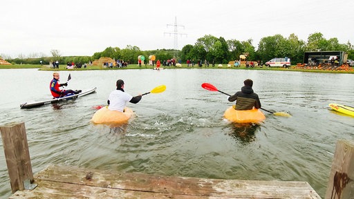 Zwei Männer sitzen in Riesenkürbissen und paddeln über den See.