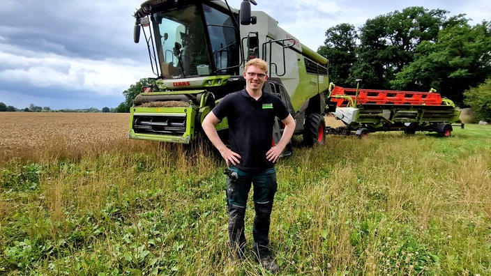 Agrar-Influencer "Klaas mit K" steht auf einem Feld und lächelt. Hinter ihm stehen große Landwirtschaftsmaschinen.