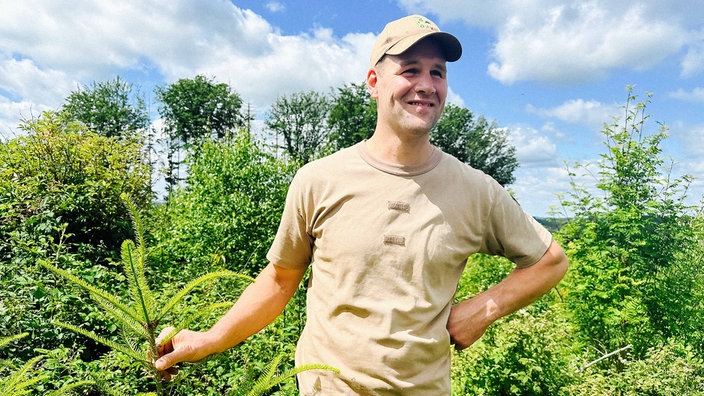 Der Landwirt und Waldbesitzer Torsten Dörmbach steht auf seinem Waldgrunstück. Er ist umgeben von Bäumen und Pflanzen und lächelt zufrieden.