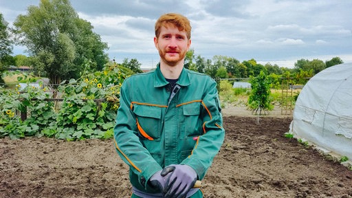 Miguel Mosdzen steht in einem grünen Overall auf dem Feld des Bochumer Tafelgartens. Mit beiden Händen stützt er sich auf einen Spaten. 