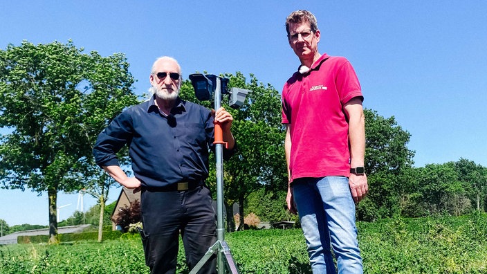 Zwei Männer stehen mit einem Bewegungsmelder auf einem grünen Feld.