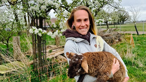 Eine Frau mit dunkelen Haaren lächelt in Richtung Kamera. Sie hält ein braunes Schaf auf dem Arm