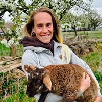 Eine Frau mit dunkelen Haaren lächelt in Richtung Kamera. Sie hält ein braunes Schaf auf dem Arm