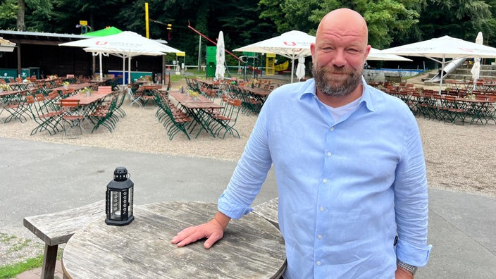 Ein Mann steht an einem Stehtisch in einem Biergarten und schaut in die Kamera.