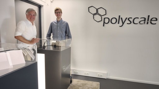 Auszubildender Simon Nobis und Geschäftsführer Tobias Wersig stehen an einer Glasvitrine und lächeln in die Kamera.