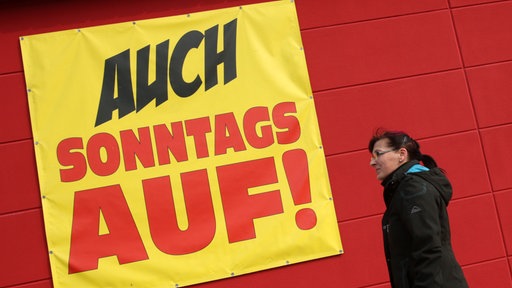 Frau läuft an Banner mit der Aufschrift "Auch Sonntags auf" vorbei