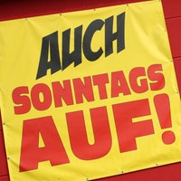 Frau läuft an Banner mit der Aufschrift "Auch Sonntags auf" vorbei