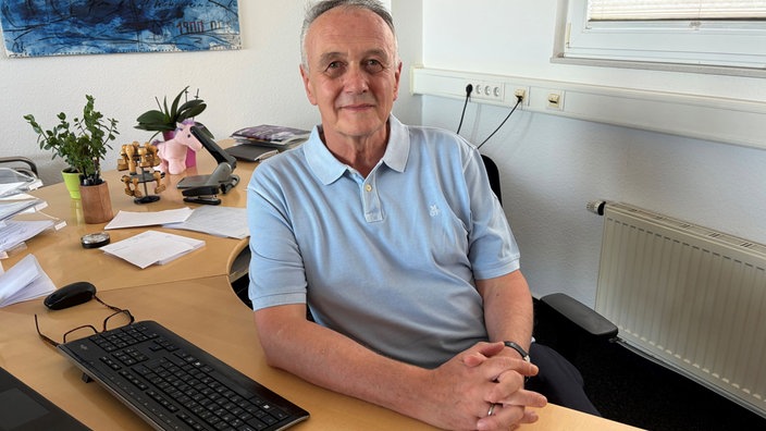 Hans Jörg Depel sitzt an einem Schreibtisch und hat die Arme und Hände darauf abgelegt, seine Hände sind gefaltet, er trägt ein hellblaues Polo-Shirt