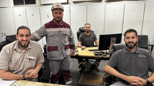 Vier Männer lächeln in die Kamera. Drei davon sitzen an Tischen und Emre Cukuk steht in seiner Arbeitskleidung dazwischen.