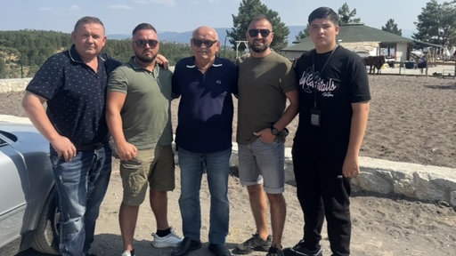 Fünf Männer schauen in die Kamera. Sie alle tragen dunkle Kleidung, drei von ihnen tragen eine Sonnenbrille. Im Hintergrund Natur und Berge.