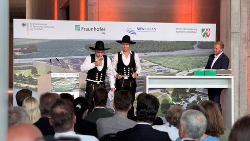 Zwei Männer auf der Bühne, während einer von ihnen Wein trinkt.