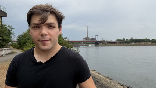 Portrait eines jungen Mannes, der am Rhein steht. Im Hintergrund der Fluss und eine Brücke.
