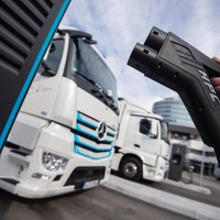 Eine Lade-Zapfhahn wird im Vordergrund von E-LKW hochgehalten.