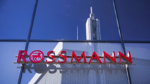 Der Rossmannschriftzug. Im Hintergrund spiegelt sich ein großes Gebäude in einem Fenster.