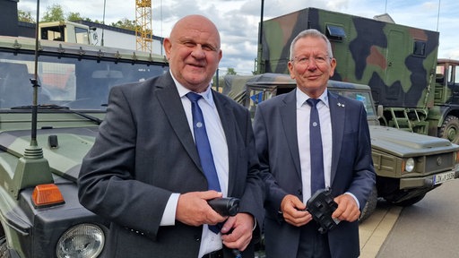 Zwei Männer stehen vor Militärfahrzeugen und halten technische Bauteile in der Hand.