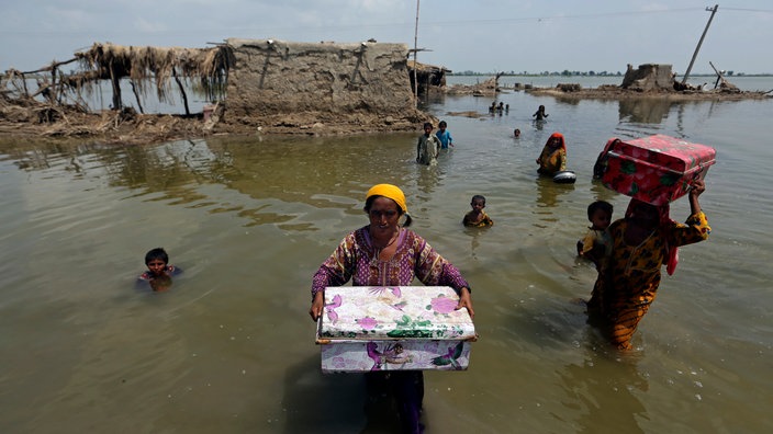 Frauen und Kinder stehen bei der Flutkatastrophe 2022 in Pakistan in hohem Wasser, sie tragen ihre übriggebliebenen Habseligkeiten