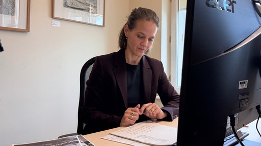 Eine Frau in einem schwarzen Blazer sitzt am Schreibtisch und liest Unterlagen, im Vordergrund ein Computerbildschirm