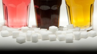 Drei Gläser gefüllt mit roter Limonade, Cola und einem Energy-Drink stehen neben Zuckerwürfeln auf einem Tisch.